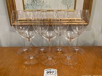 Eight clear Riedel white wine glasses arranged in two rows on a wooden surface in front of a framed artwork.