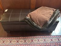 Side view of dark brown faux leather ottoman with brown knit throw draped on top near wooden panel wall and patterned rug