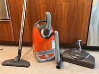 Full front view of orange vacuum with hose, wand and two floor heads