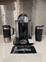 Front view of Nespresso machine with ribbed water tank, two travel mugs and Aeroccino3 manual on countertop