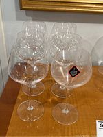 Six clear Riedel red wine glasses arranged in two rows on a wood surface with a framed picture behind them. One glass shows the original Riedel sticker.
