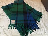 Full view of the folded blue and green tartan wool blanket with fringed edges on a bed.