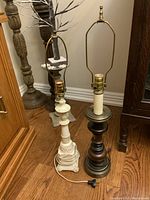 Two table lamps side by side on a wooden floor, one with a white ornate ceramic base and brass top, the other with a dark wooden turned base and brass top. No lampshades shown.