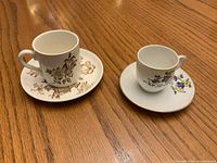Front view of two miniature ceramic cups with matching saucers showing floral patterns in brown and multicolor.