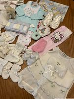 Photo of baby clothing and accessories spread on table showing multiple pairs of booties, onesies, socks, mittens, bib, and blanket