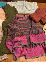 Nine hand knit sweaters, poncho, and vest in various colors and knitting patterns, displayed together on a wooden surface.