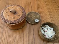 Photo showing three woven baskets: one medium brown basket with a stitched leather-like lid and handle, a small round basket with buttons and sewing items, and a small basket lid with a pin.