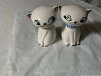 Pair of ceramic white cat salt and pepper shakers with green eyes, one with a blue collar and one with a pink collar, placed on white crinkled background.