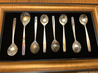 Set of 8 silver spoons arranged side-by-side for display, showing tarnished surfaces and varied spoon sizes.