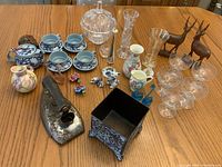 Photo of miscellaneous decorative and tableware items on wooden surface including porcelain, glassware, metal figurines, and ceramics.