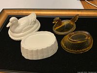 Photo showing white ceramic duck covered dish with lid and matching base, amber glass rooster covered dish with lid and separate base, all arranged on black background.