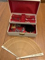 Open vintage jewelry box showing red fabric interior with compartments filled with assorted sterling and costume jewelry, two old wristwatches, black accessory, and carved wooden fan placed in front.