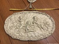 Frontal photo of the oval wall plaque showing three cherubs in relief with a measuring tape for size reference horizontally.