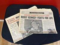 Two vintage newspapers stacked, front page visible with headline about Bobby Kennedy critically wounded, dated June 5, 1968.