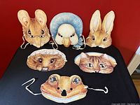 Front view of seven Beatrix Potter character masks arranged on black surface, showing detailed illustrations of Mr Jeremy Fisher Frog, Tom Kitten, Mrs Tiggy Winkle, Jemima Puddle-Duck, The Tailor of Gloucester Mouse, rabbit and hedgehog faces.