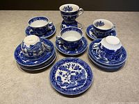 Blue Willow china tea cups and saucers arranged with their corresponding plates, showing the bottom markings on some cups and the intricate blue landscape designs on the items.