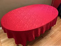 Red oval-shaped tablecloth with brocade rose pattern displayed on wooden floor, showing full coverage and texture.