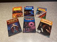 Photo of all six Galaxy Science Fiction magazines laid out on a flat surface showing front covers with distinct colorful space-themed artwork from mid-1970s.