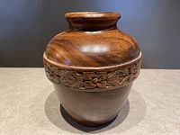 Front and angled view of hand turned wooden vase with carved floral band around upper section, showing detail of wood grain and floral carving.