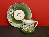 Front view of the teacup with painted flowers on a green background, showing the handle and Cyrillic script, with the matching saucer behind