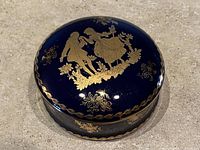 Top view of round porcelain trinket dish with deep blue glaze and detailed gold illustration of a couple in an outdoor scene surrounded by floral motifs.