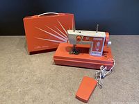 Orange and white Little Queen portable toy sewing machine shown with foot pedal and original carrying case. Machine has tension knob, stitch selector dial, spool pin and needle bar visible.