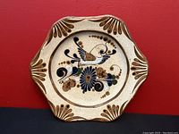 Octagonal ceramic wall plate with folk art bird design against a cream speckled background and scalloped brown edges, hanging against a red wall.