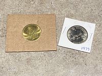 Two coins side by side on concrete: gold-colored 2002 coin on left in cardboard holder, silver-colored 1979 coin on right in white holder labeled '1979'.