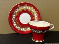 Front-facing photo of the Burgundy red teacup with gold and white filigree design, placed upright next to matching saucer against a beige background.