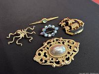 Five vintage brooches arranged on a black background: gold-tone octopus, ornate brooch with pearl cabochon, rhinestone oval brooch, tie pin with green gem, and ship brooch with enamel and gems.