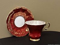 Side view of the red and gold Royal Albert teacup with matching saucer behind it, showing intricate gold details and scalloped edges.