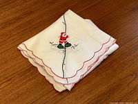 Folded vintage white Christmas tablecloth with embroidered Santa and scalloped red edges.