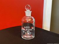 Clear vintage glass apothecary jar with stopper lid against red background showing front label Acid Sulfuric H2SO4.