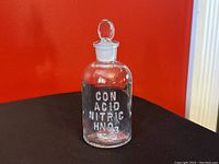 Front angle of vintage clear glass apothecary jar labeled CON ACID NITRIC HNO3 with lid against red background