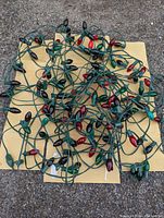 Photo showing the tangled string of red and green large glass bulb Christmas lights on green wire against a yellow and concrete background.