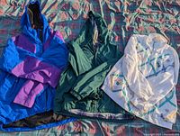 Three ladies jackets laid out flat: blue and purple hooded jacket, green hooded jacket, and white jacket with design.