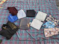 Overview photo of 11 pieces of men's clothing laid flat on a plaid blanket showing sweaters, pants, a jacket, and button-up shirts in various colors.