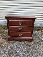 Night end table front view showing wooden dresser with three drawers and brass handles, standing outdoors on gravel in front of a light-colored wall.