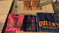 Two Queens University sweaters, one pink and one navy blue, a Queens themed multicolored plaid scarf, and a pair of wooden bookends with metal plaques showing university crests placed on cardboard outdoors.
