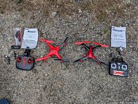 Two red and black quadcopter drones with their controllers, batteries, manuals, extra propeller blades, and cables arranged on the ground.