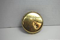 Gold-tone jar/bottle opener showing 'lige til ollet' inscription on top, round shape with serrated edges.