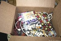 Top-down photo of an open cardboard box filled with assorted jewelry items mostly necklaces, bracelets and crafting materials.