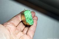 Hand-held view of green quartz stone pendant showing gold paint side and metal loop attachment.