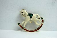 Front close-up photo of a small enameled rocking horse brooch showing cream, green, red and gold-tone details.