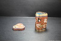 Photo showing cylindrical body and separate heart-shaped lid placed on a flat surface, showcasing enamel design and metal edging.