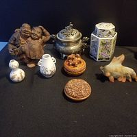 Display of entire lot including wooden figurines, porcelain vases, metal container, decorative jars, and carved wooden and resin items arranged on black background.