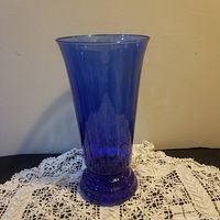 Front view of a tall cobalt blue hand-blown vase sitting on a lace tablecloth against a plain wall.