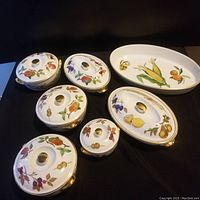 Six pieces including five covered serving dishes and one platter on black background showcasing shapes, motifs, and gold trim