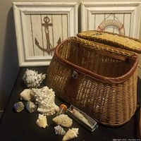 Wicker fishing creel basket with assorted seashells in front and two nautical-themed framed prints leaning against the wall.