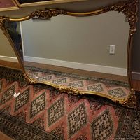 Full view of a large rectangular mirror resting on the floor, showing its ornate decorative gold frame and reflective glass surface.
